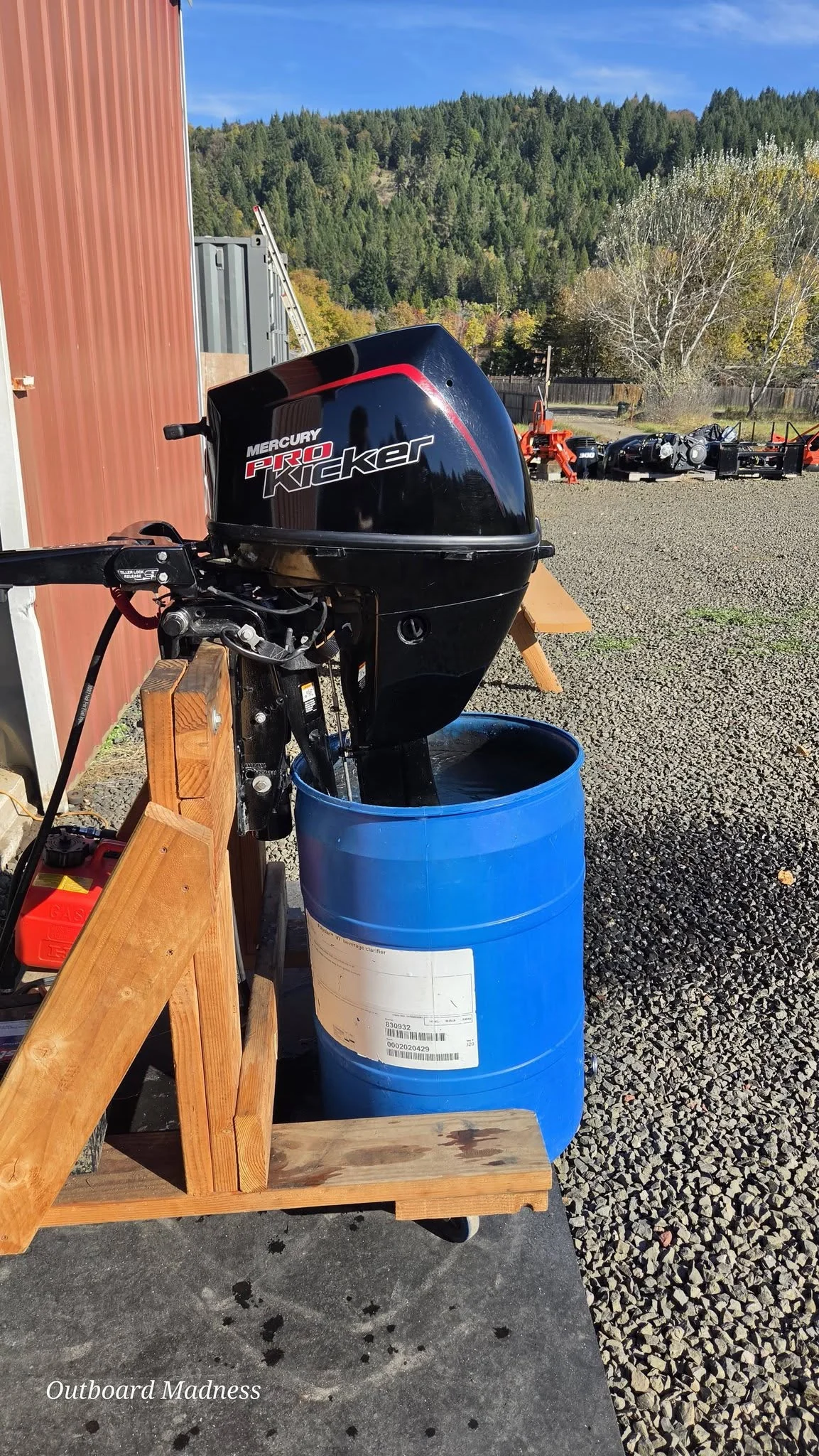 A black Mercury outboard clamped to a wooden stand, its lower unit in a blue water barrel beside the red workshop wall, with a wooded hillside behind.